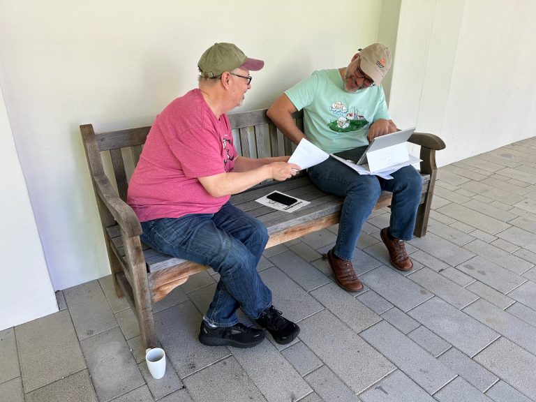 Joe Yoder and Christian Köppe with caps on and laptop open, sitting at a bench engaged in conversation