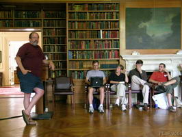 Ward Cunningham dressed in casual shorts speaking to four people sitting by a bookshelf-lined wall